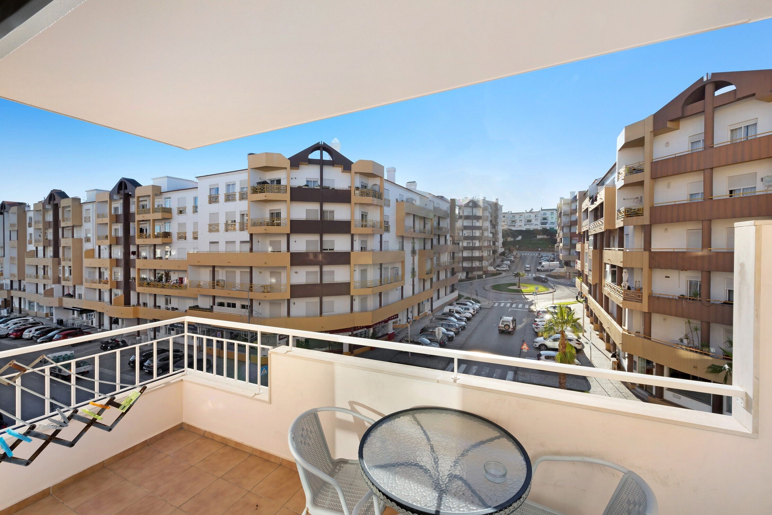 A balcony with a glass table and chairs, overlooking a street with parked cars and multi-story buildings.
