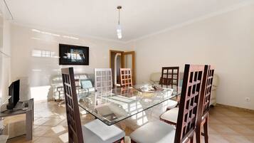 A dining area with a glass-top table, wooden chairs, a television, and a tiled floor.