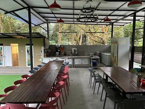 An outdoor dining area with a long wooden table and red chairs, a kitchen area with cooking equipment, and a view of trees and greenery. - Private Pool FamilyResort Bentong Pahang (Raub)