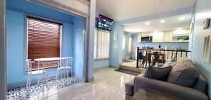 A living room with a sofa, a TV mounted on the wall, and a kitchen area in the background.
