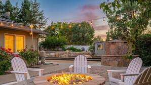 A patio with a stone fire pit surrounded by white Adirondack chairs, string lights, and a stone wall. - 4,500 sq-ft retreat with a pool and hot tub in the middle of wine country!  (Templeton)