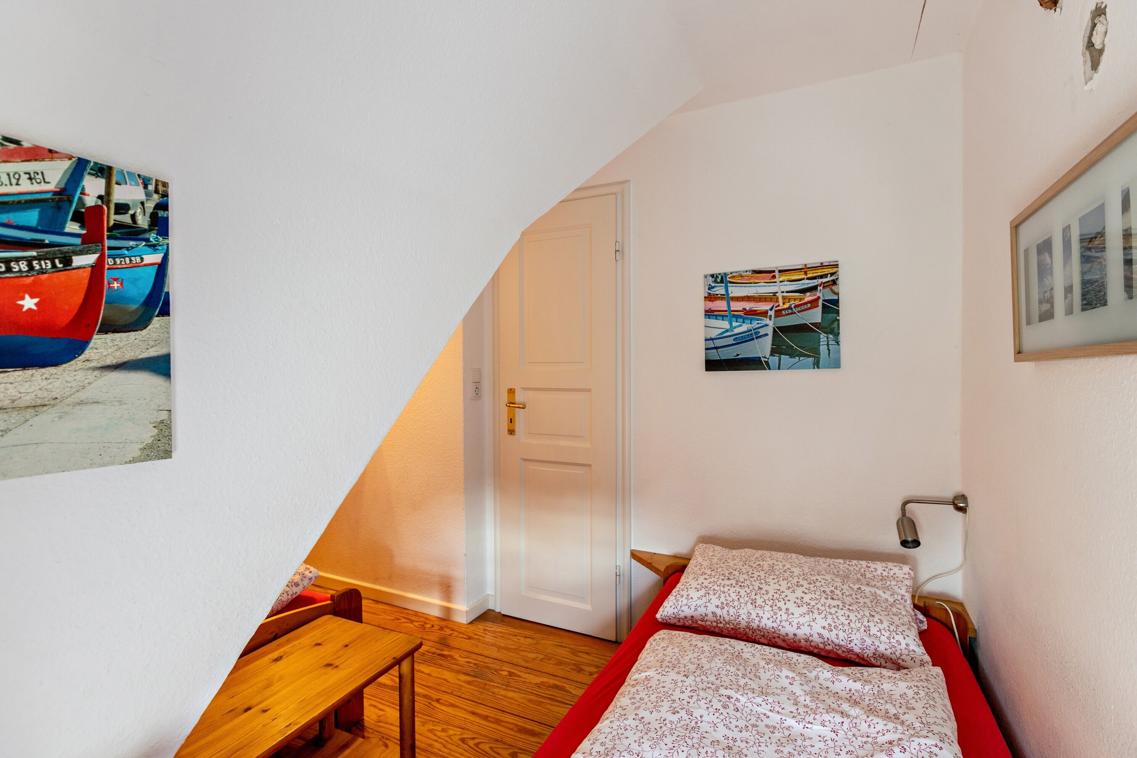 Une pièce avec un banc en bois, un lit recouvert d’une literie à motifs floraux, une porte blanche et deux tableaux encadrés accrochés au mur.