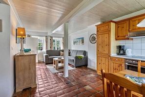 A cozy living room with a sofa, a coffee table, and a dining table with chairs. There is a kitchen area with wooden cabinets and a stove.