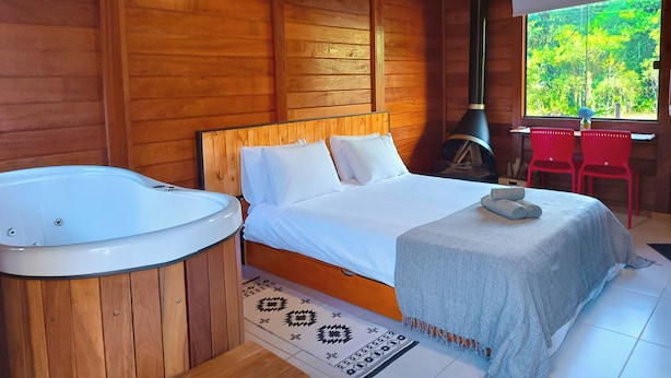 Habitación de cabaña de madera con cama, bañera de hidromasaje y ventana con vistas a la vegetación.