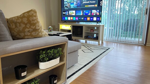 A modern living room with a flat-screen TV displaying a streaming service interface, a grey sofa, a wooden coffee table, and a large window with blinds. - Comfy 2 bedroom condo with 75” smart TV (Chicago Ridge)