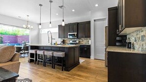 A modern kitchen with dark cabinets, a central island with bar stools, pendant lighting, and a stone backsplash. - Ivins Oasis : Private Hot Tub & Pool (Ivins)