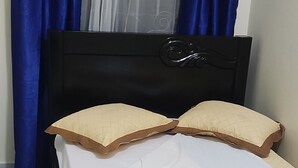 Una cama con cubierta marrón acolchada, dos almohadas beige y un sábana blanca. Se ven cortinas azules y una cortina translúcida al fondo.