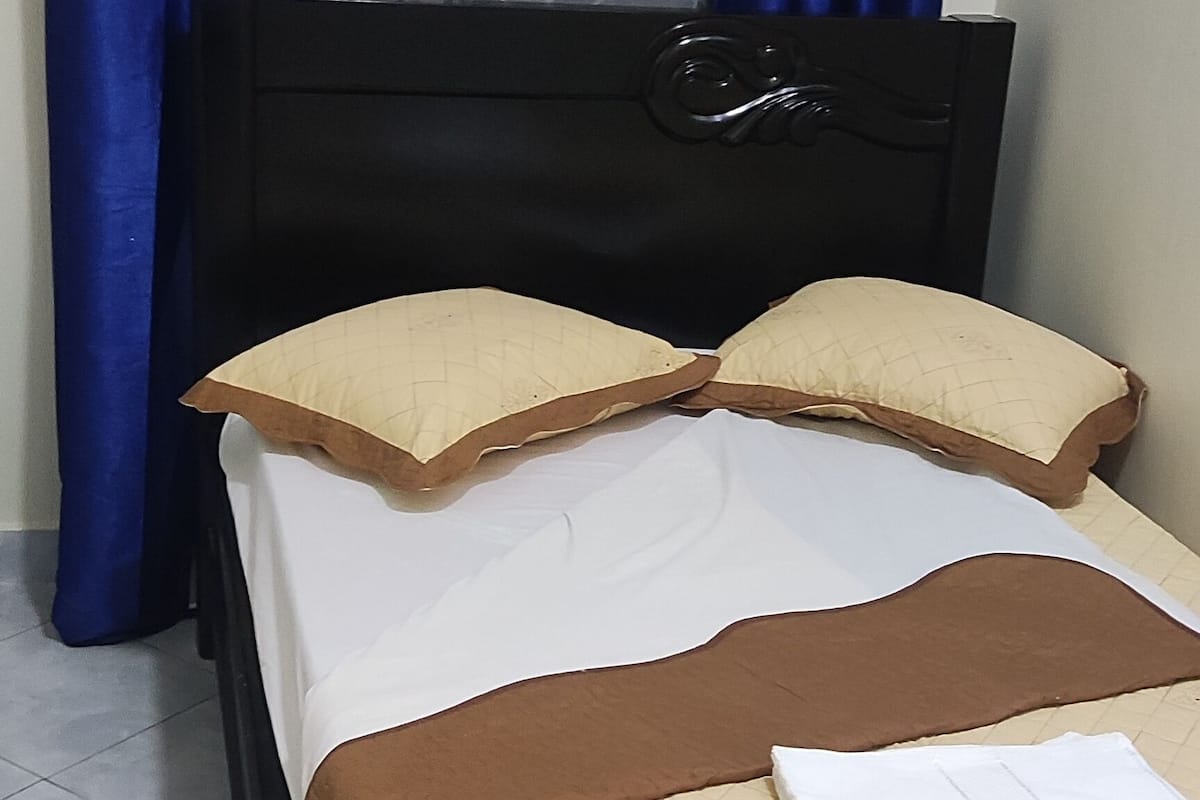 A bed with a quilted brown cover, two beige pillows, and a white bedsheet. Blue curtains and a sheer curtain are visible in the background.
