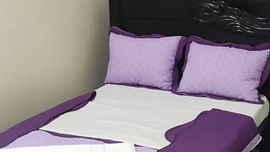 Una cama con una colcha morada y blanca, un cabecero negro y una ventana con cortinas azules.