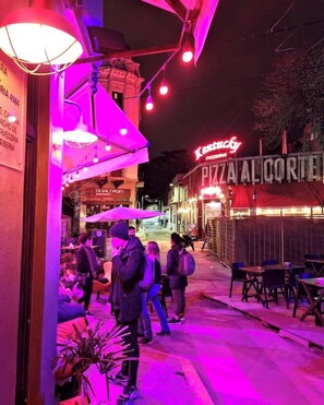 Une scène de rue avec un restaurant de pizzas appelé « Pizza Al Cortez » et un bâtiment sur lequel on peut lire l’inscription « Dura Vida ».