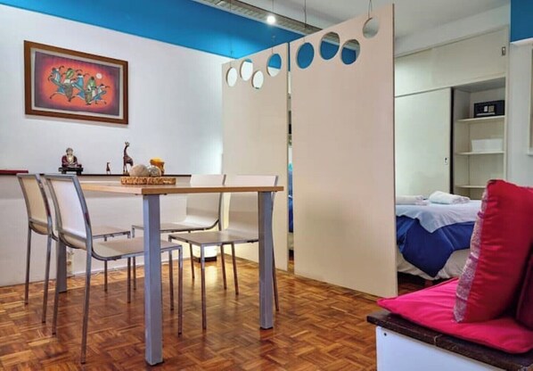 Une pièce avec un plancher en bois, une table à manger avec des chaises, un lit avec un oreiller rouge et un mur orné d’un tableau encadré.