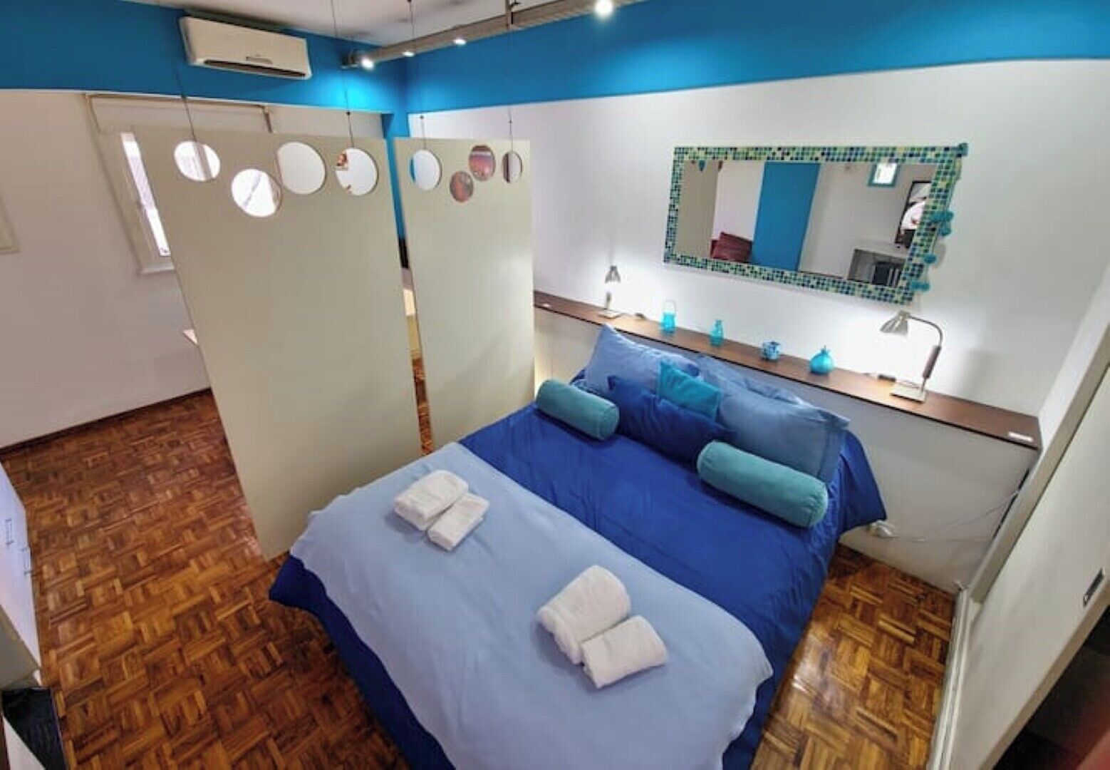 A bedroom with a blue bed, two white towels, a mirror with a green frame, and a wooden floor.