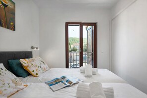 A hotel room with a bed, pillows, a magazine, a balcony with a view of greenery and buildings.