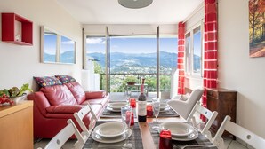 A dining area with a table set for two, a red leather sofa, and a view of mountains through large windows. - Pazzallo Panoramica with Balcony and Garage, Lugano, Switzerland (Lugano)