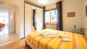 A hotel room with a large bed, a bedside table with a bottle and glasses, a window with curtains, and a hallway leading to another room. - Pazzallo Panoramica with Balcony and Garage, Lugano, Switzerland (Lugano)