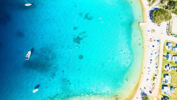 Une vue aérienne d’une plage aux eaux turquoise et limpides, avec quelques bateaux et une rangée de petits chalets situés en bord de mer.