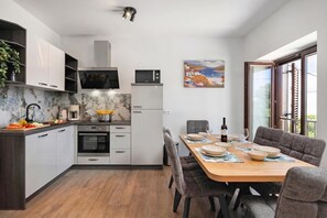 A modern kitchen with white cabinets, a marble backsplash, and a dining area with a wooden table and grey upholstered chairs. - Seafront Apartment Nana 1, Punat, Croatia (Punat)
