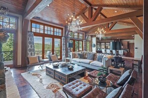 A spacious living area with a fireplace, large windows, and a high ceiling with exposed beams. - Alpine Sanctuary | Walk-to-Ski Access | Private Hot Tub | Media & Game Room (Park City)