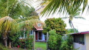 Une maison dotée d’un porche à l’avant, entourée d’une végétation luxuriante et de palmiers.