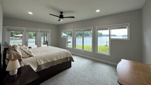 Un dormitorio con una cama grande, ventilador de techo y vista a un lago a través de amplios ventanales.