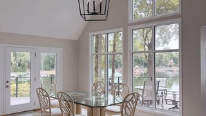 Un comedor con mesa de vidrio y cuatro sillas, ventanas amplias con vista a árboles y una lámpara colgante.