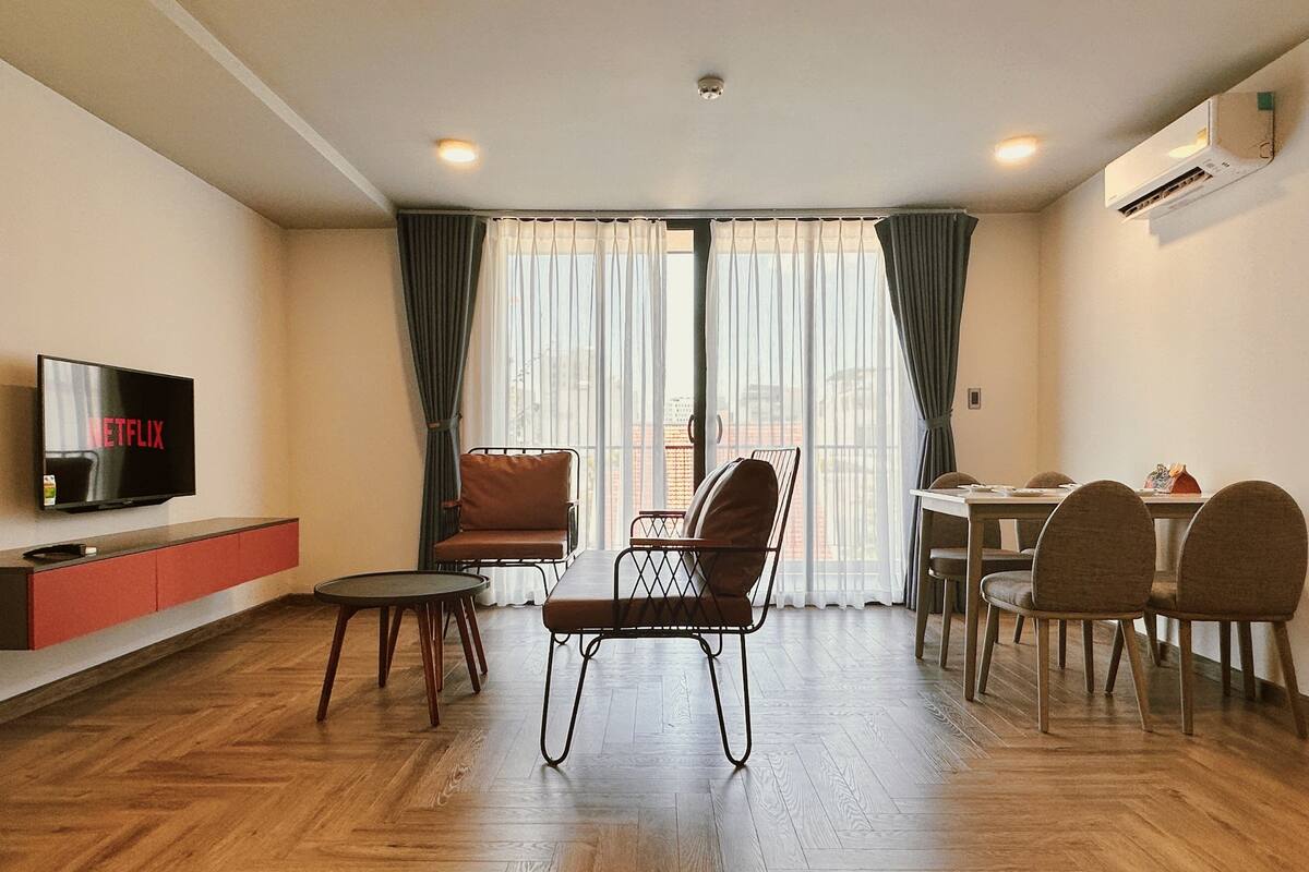 Ein modernes Wohnzimmer mit Flachbildfernseher, einer Couch, einem Esstisch mit Stühlen und einem großen Fenster mit Vorhängen.