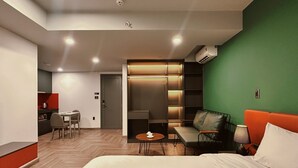 A modern hotel room with a large bed, a green accent wall, a seating area with a sofa and armchair, a small table with a lamp, and a kitchenette in the background. - TUK TAK SPACES (Da Nang)