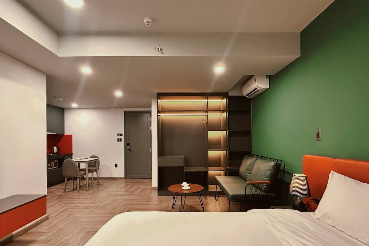 Ein modernes Hotelzimmer mit einem großen Bett, einer Wand in Grüntönen, einer Sitzecke mit Sofa und Sesseln, einem kleinen Tisch mit Lampe und einer Küchenzeile im Hintergrund.