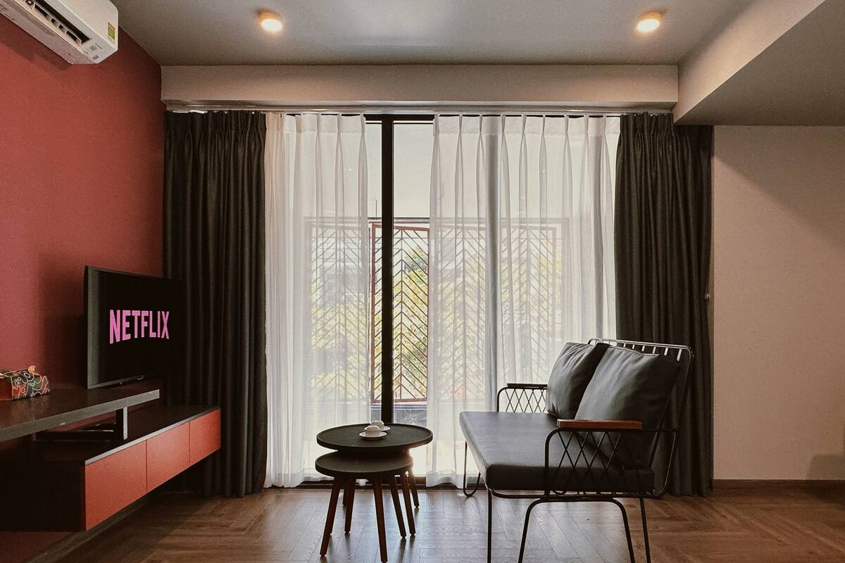 Ein modernes Wohnzimmer mit einem Flachbildfernseher, der 'NETFLIX' anzeigt, einer Couch, einem kleinen runden Tisch und einem Balkon mit Vorhängen.