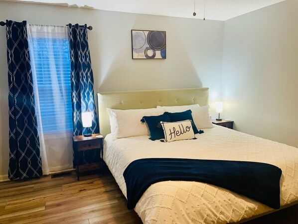 A bedroom with a bed, a nightstand, a bedside lamp, a window with curtains, and a wall art piece.