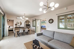 A modern living room with a grey sofa, a wooden coffee table, and a dining area with a table and chairs. There is a large window with a view of the outside.