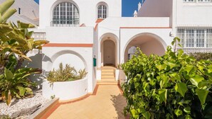 Um edifício branco com arcadas e janelas, um caminho de azulejos e plantas verdes.