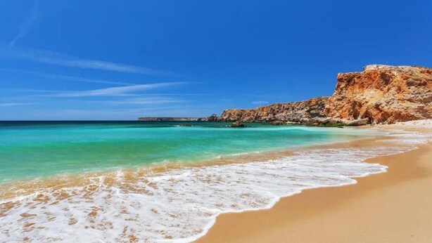 Uma praia com águas turquesa cristalinas, areal arenoso e um escarpe rochoso.