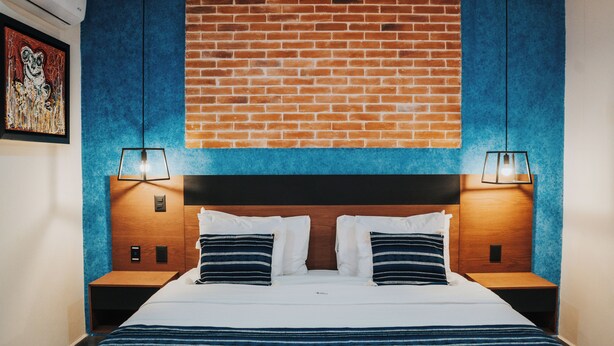 A hotel room with a brick wall, a large bed with striped bedding, two bedside tables with lamps, and a framed picture on the wall. - Hotel Casa Puga (Oaxaca)