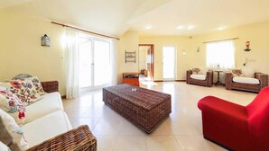 A spacious living room with a large window, a red sofa, wicker furniture, and a wooden coffee table. - Villa Brejos in Albufeira (Albufeira)