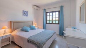 A bedroom with a bed, bedside tables, a desk, a window with curtains, and an air conditioning unit. - Villa Quinta do Jolu (Albufeira)