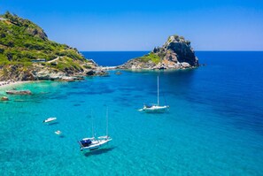 A coastal landscape with clear turquoise waters, rocky outcrops, and a few sailboats anchored near the shore.