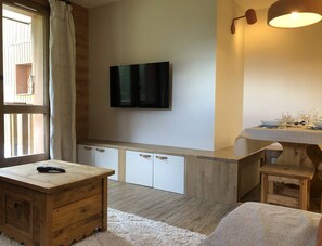 Een ruimtelijk ingerichte leefruimte met een aan de muur bevestigde televisie, een houten kast met lades, een tafeltje en een raam met gordijnen.