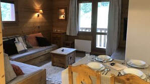 A cozy cabin interior with a wooden sofa, a dining table set for two, and a large window with curtains. - Lauzière Dessus 22 - Crève Coeur (LES AVANCHERS VALMOREL)