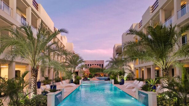 A swimming pool with lounge chairs and palm trees, surrounded by hotel buildings.