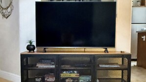 Ein Wohnzimmer mit Flachbildfernseher an der Wand, einem Holztisch im Vordergrund und einer Küchenzeile auf der rechten Seite.