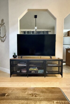 A living room with a flat-screen TV mounted on a wall, a wooden coffee table in the foreground, and a kitchen area to the right.