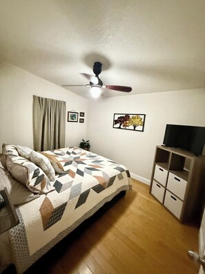 A bedroom with a bed, a ceiling fan, a TV stand, and a wall-mounted TV.