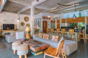 Un vaste espace de vie comprenant un canapé, une table basse en bois et des poufs en osier. Il y a également un coin cuisine avec des tabourets de bar et une table à manger.
