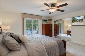 Un dormitorio con una cama grande, un ventilador de techo, una ventana con vistas, un aparador de madera y un televisor en la pared.