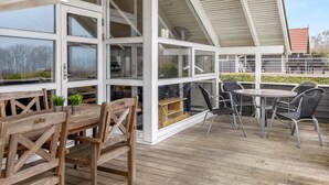 A covered outdoor area with a wooden deck, multiple tables and chairs, and large windows. - "Hera" - 200m from the sea by Interhome (Sydals)