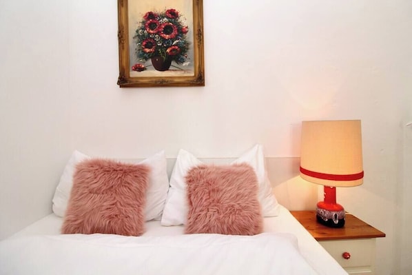 A bed with white bedding and two fluffy pink pillows, a wooden nightstand with a red lamp, and a framed painting of flowers on the wall.