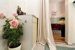 A hallway with a potted rose plant, a cabinet, and a doorway leading to a room with a patterned floor and a red sofa.