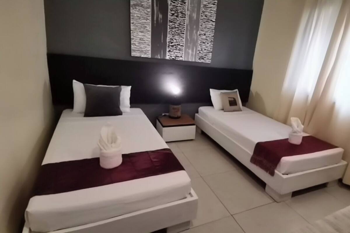 A hotel room with two beds, a nightstand, a lamp, and a window with curtains.