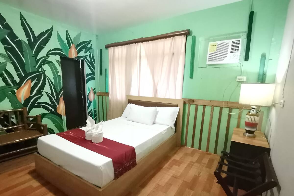 A single bed with a wooden headboard, a red blanket, and white pillows. There is a small table with a lamp and a chair to the right. The room has green walls with a large tropical leaf mural.